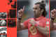 A man with long hair tied back wears a red sports jersey, raises both index fingers, and holds the shirt collar with one hand.